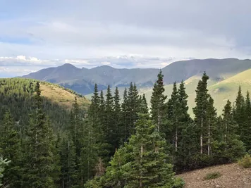 evergreen trees in the foreground with rolling mountain ranges under a cloudy sky in the background