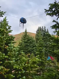 A blue and black backpack suspended on a zip line among evergreen trees with a red tent partially visible in the background